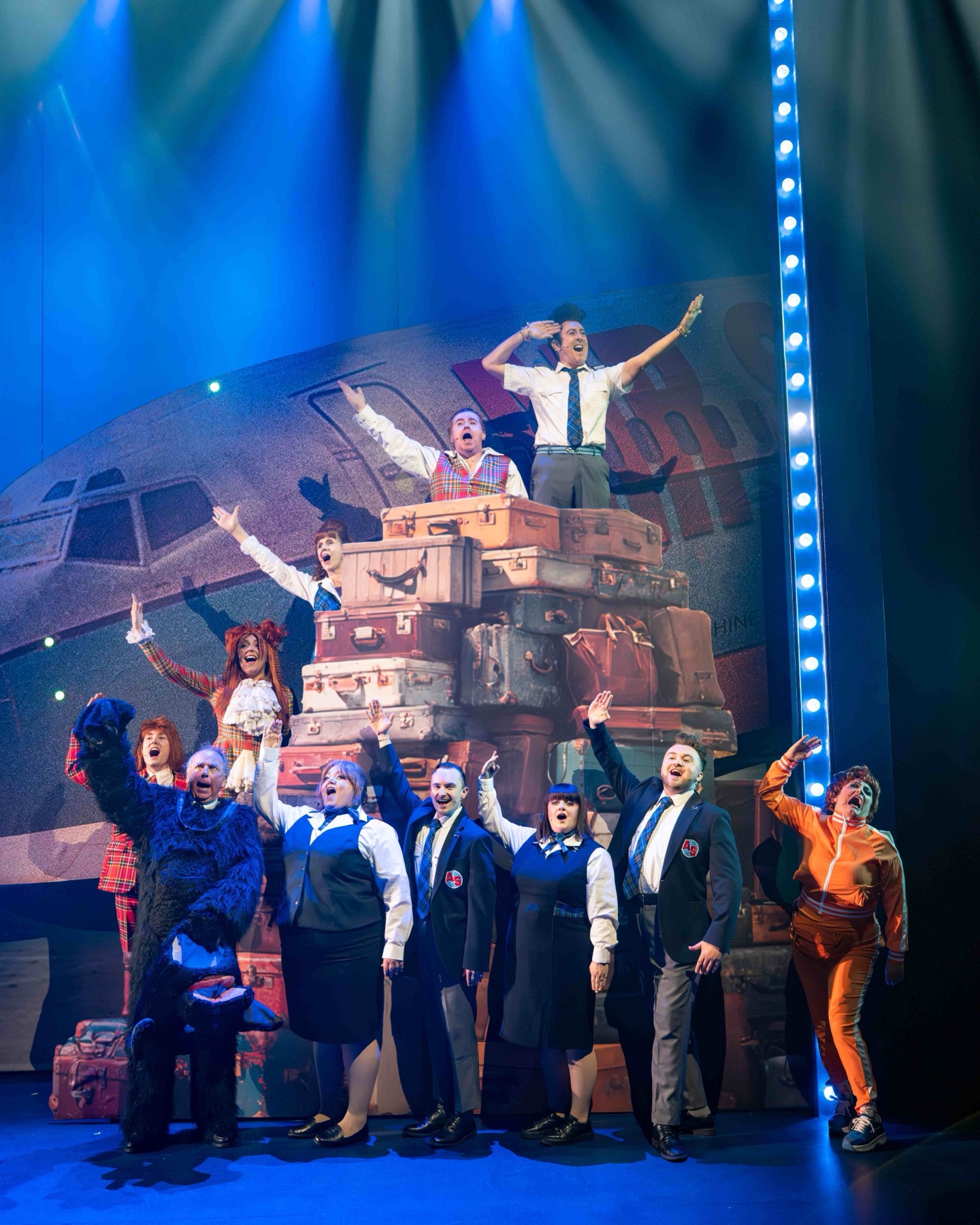 The High Life cast wave in front of a towering stack of suitcases.